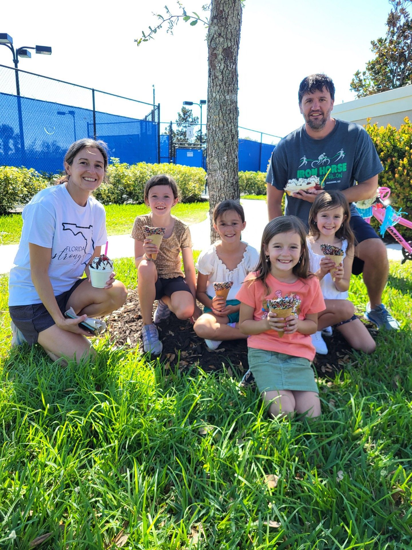 Group of adults and children smiling with ice cream cones next to a tree. Green grass, tennis courts in background.