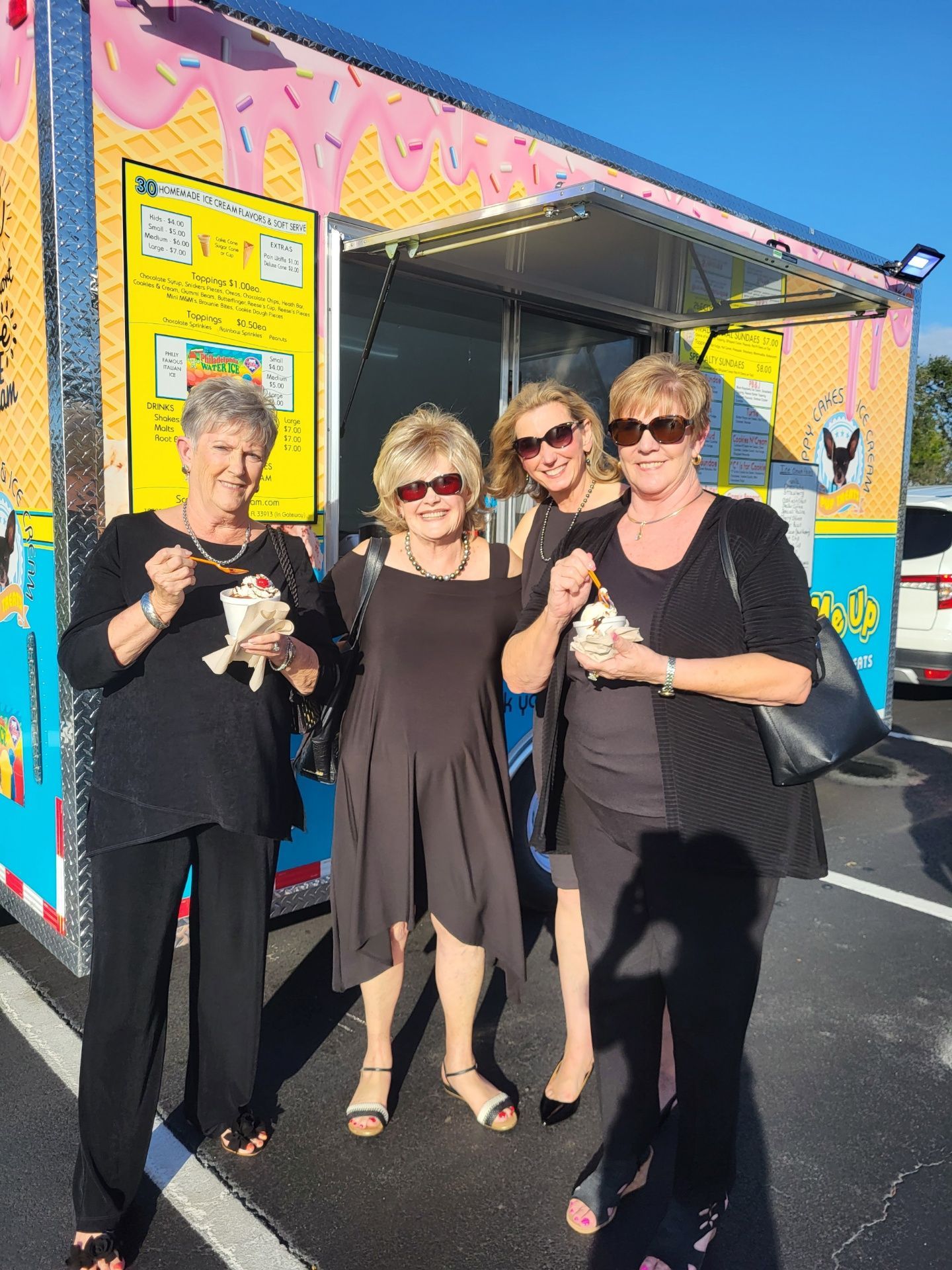 Four women in black outfits holding ice cream near an ice cream truck on a sunny day.
