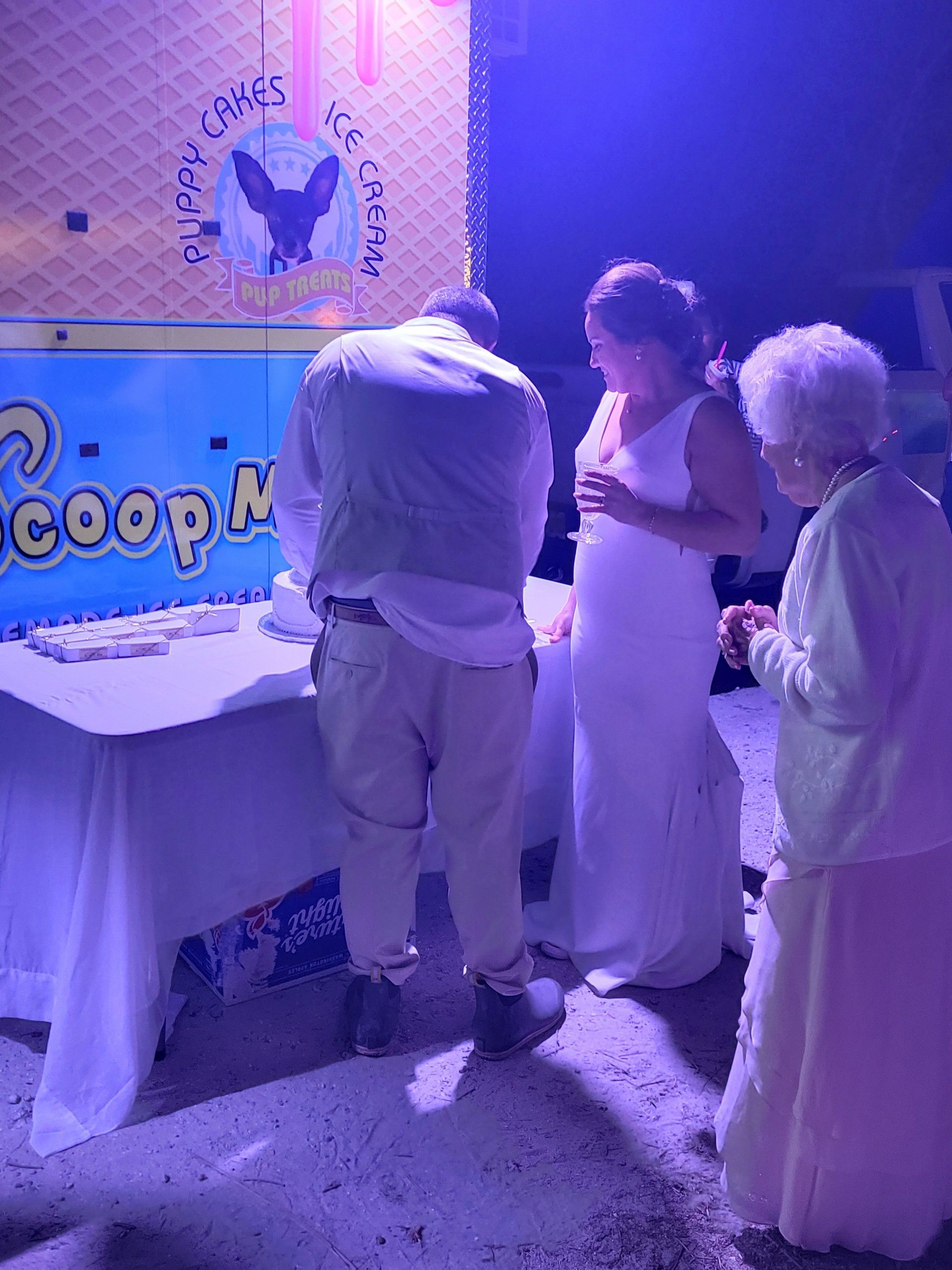People at ice cream table; man scoops, woman in white dress and elderly woman watch, at night.