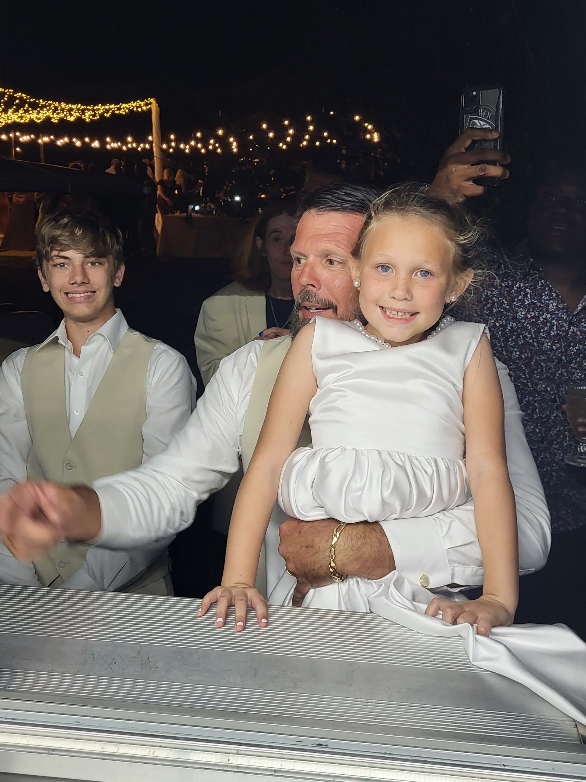 Man holding girl in white dress, a boy in a vest, and background of outdoor night event.