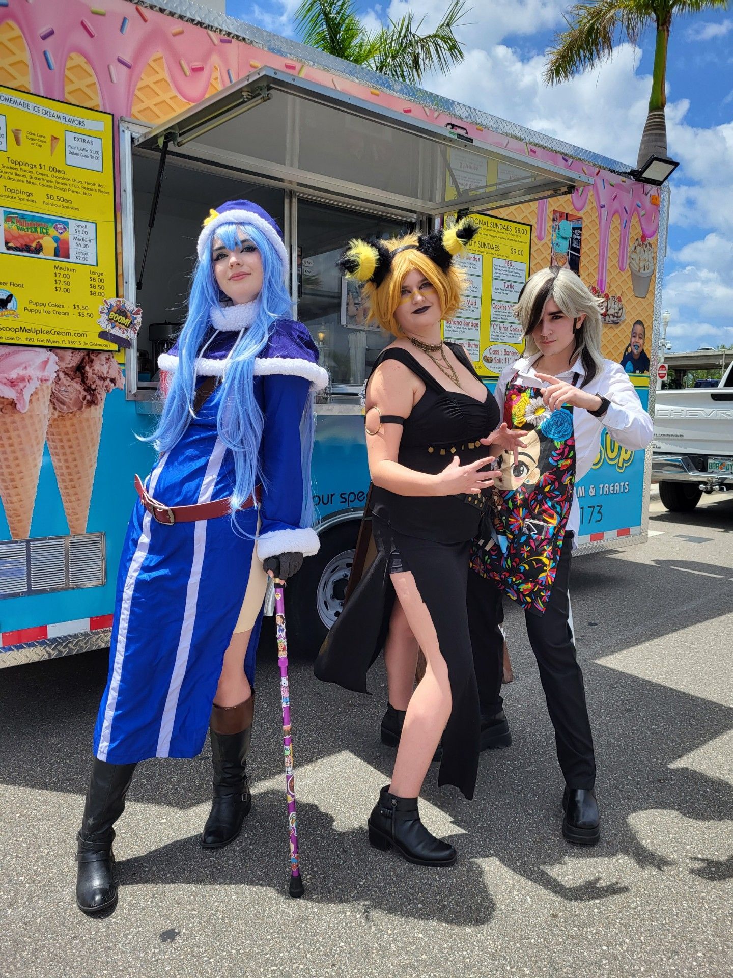 Three cosplayers pose in front of an ice cream truck. Blue-haired woman, yellow-haired woman, man in black, sunny day.