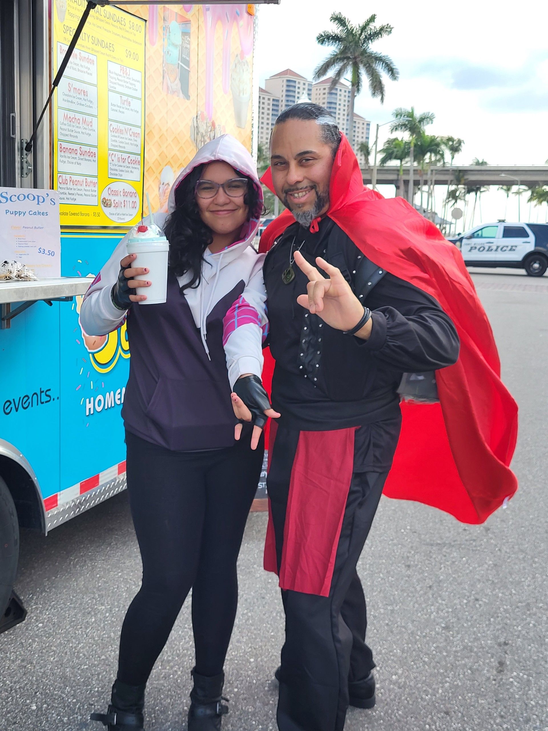 Woman and man pose near a food truck. The man wears a costume with a red cape. The woman holds a drink.