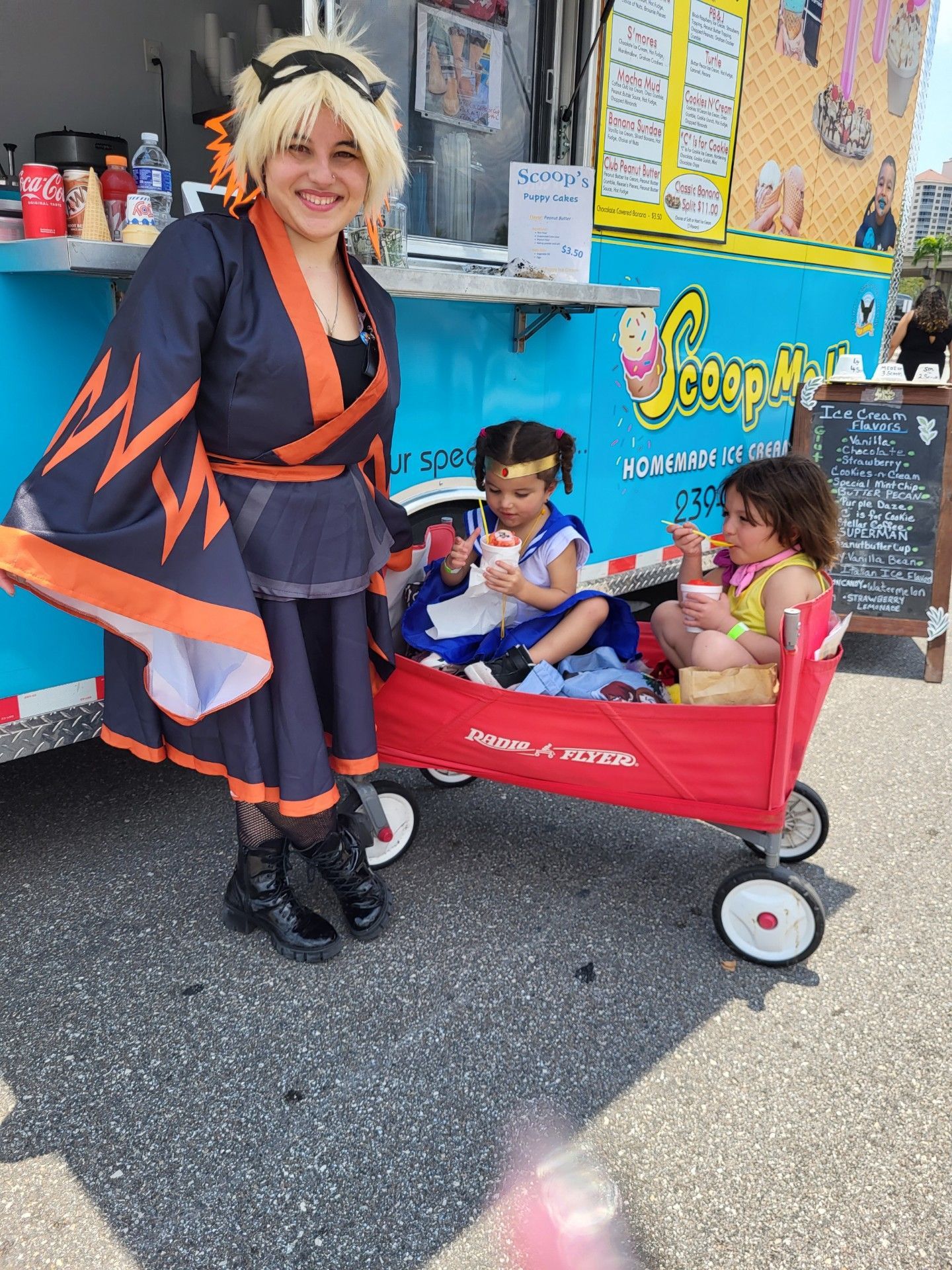 Woman in costume poses with two children in a wagon near a blue ice cream truck.