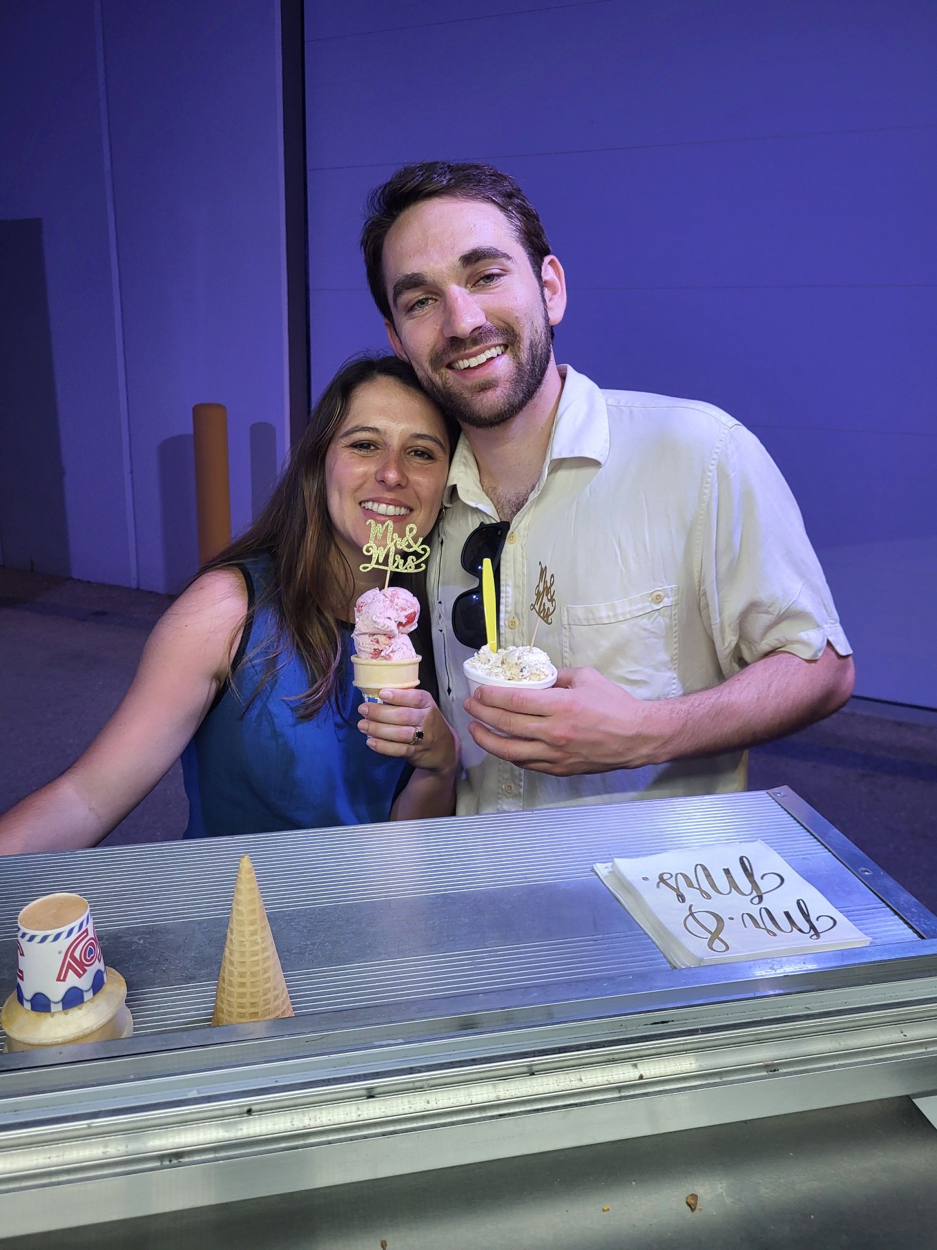 Smiling couple holding ice cream cones; woman in blue, man in beige shirt, standing next to a display.