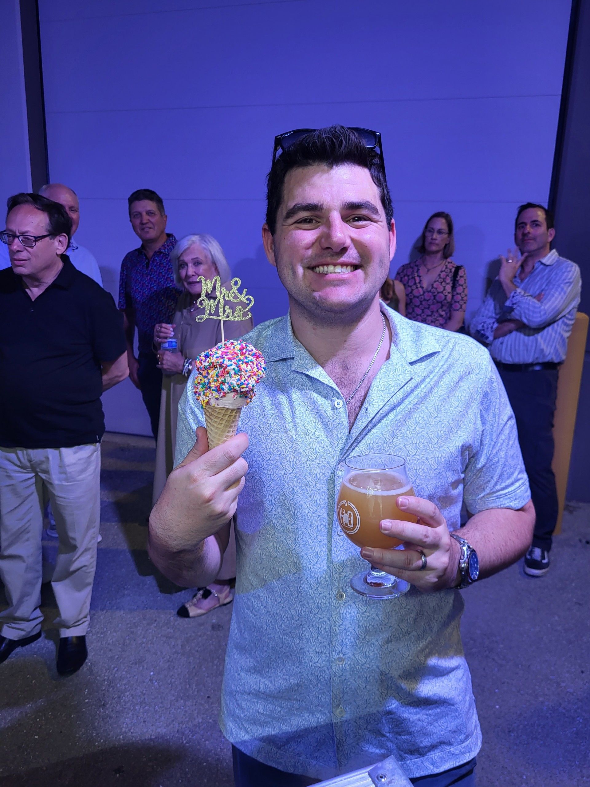 Man smiles, holds ice cream cone & beer.  People stand in a blue-lit setting.