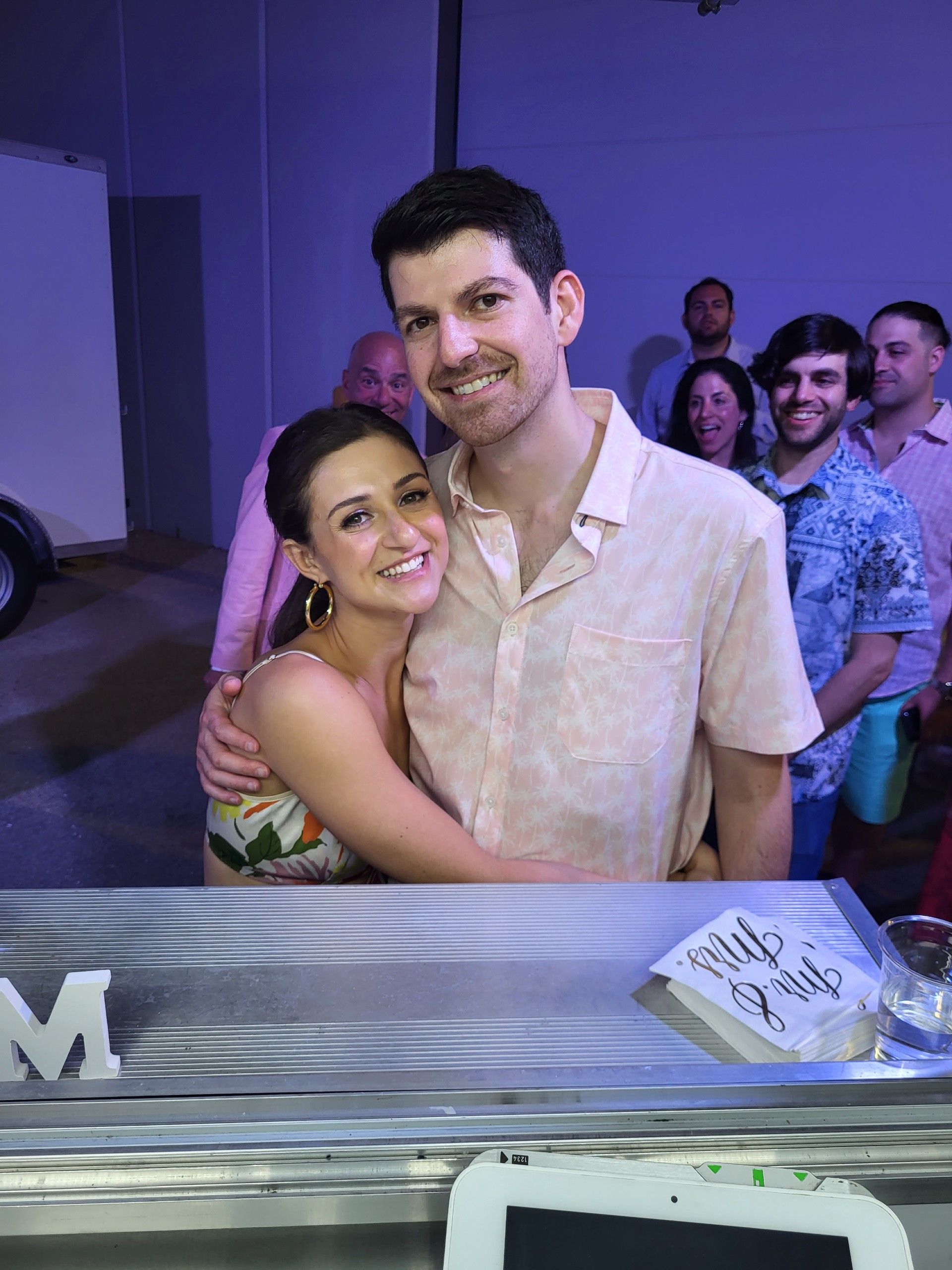 Couple embraces, smiling. Pink and floral tops, others in the background. Party or event indoors.