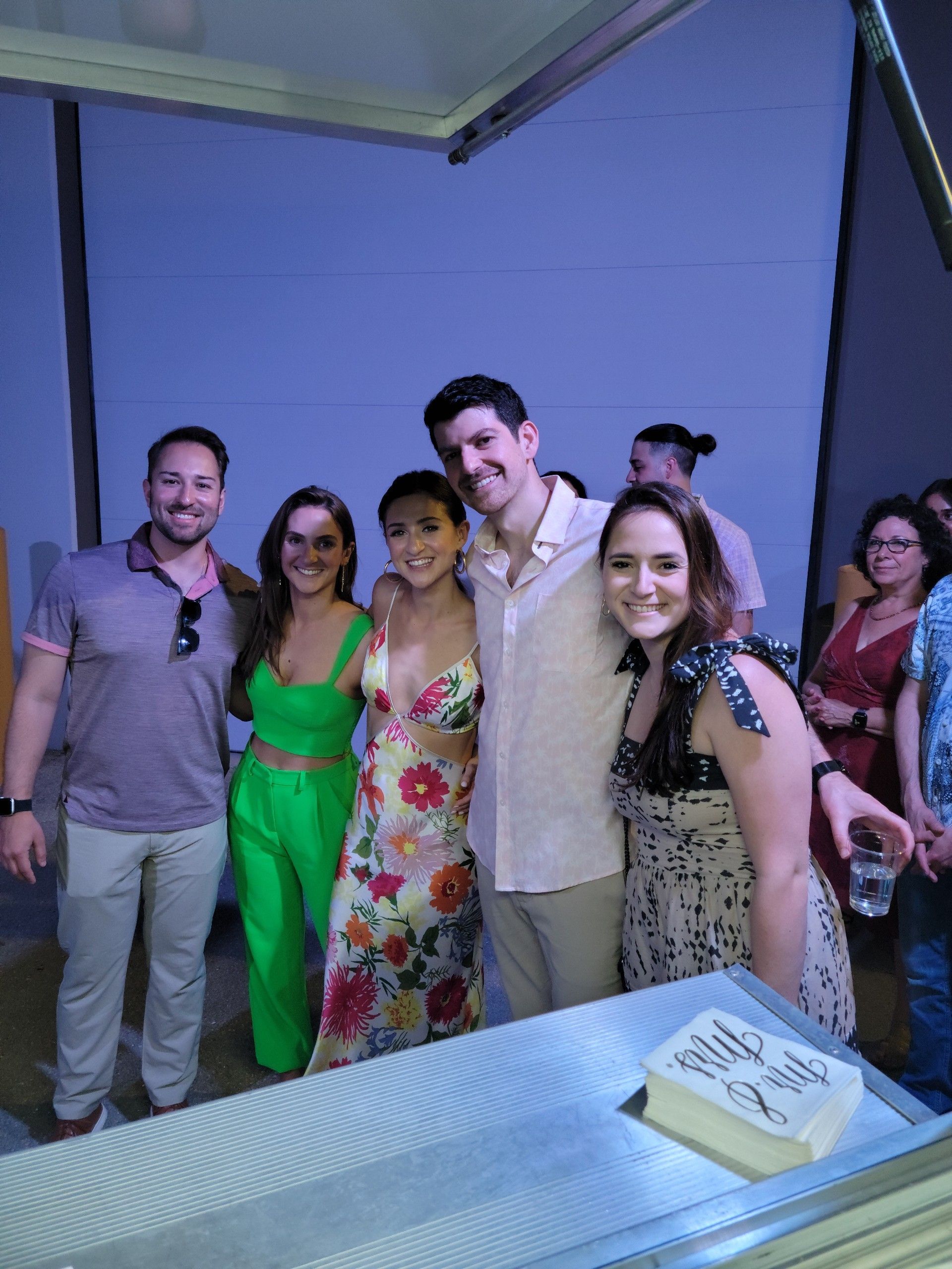 Group of people posing for a photo, a woman in a floral dress smiles. Light blue background.