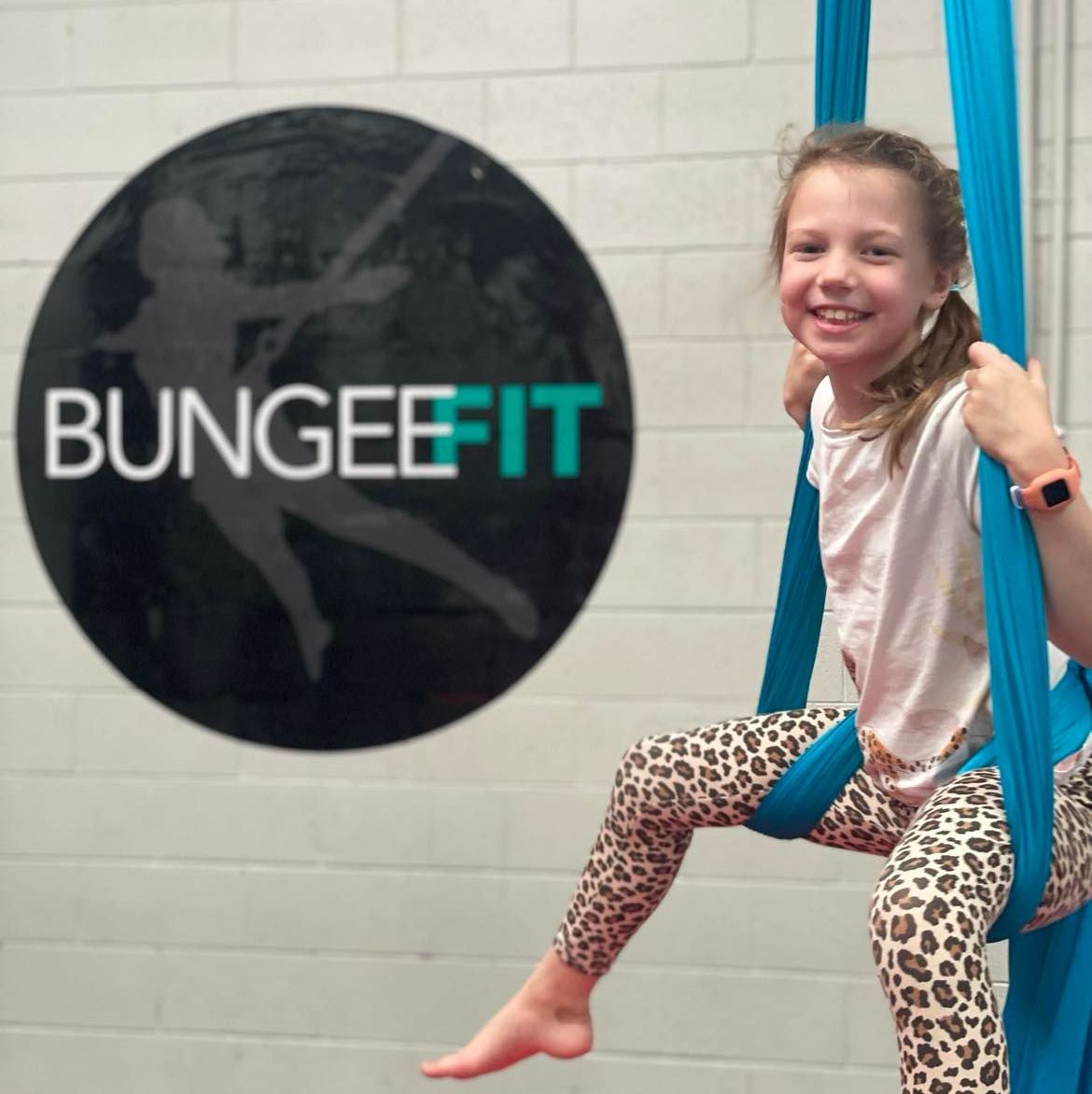 A young girl is sitting on a blue bungeefit aerial silk