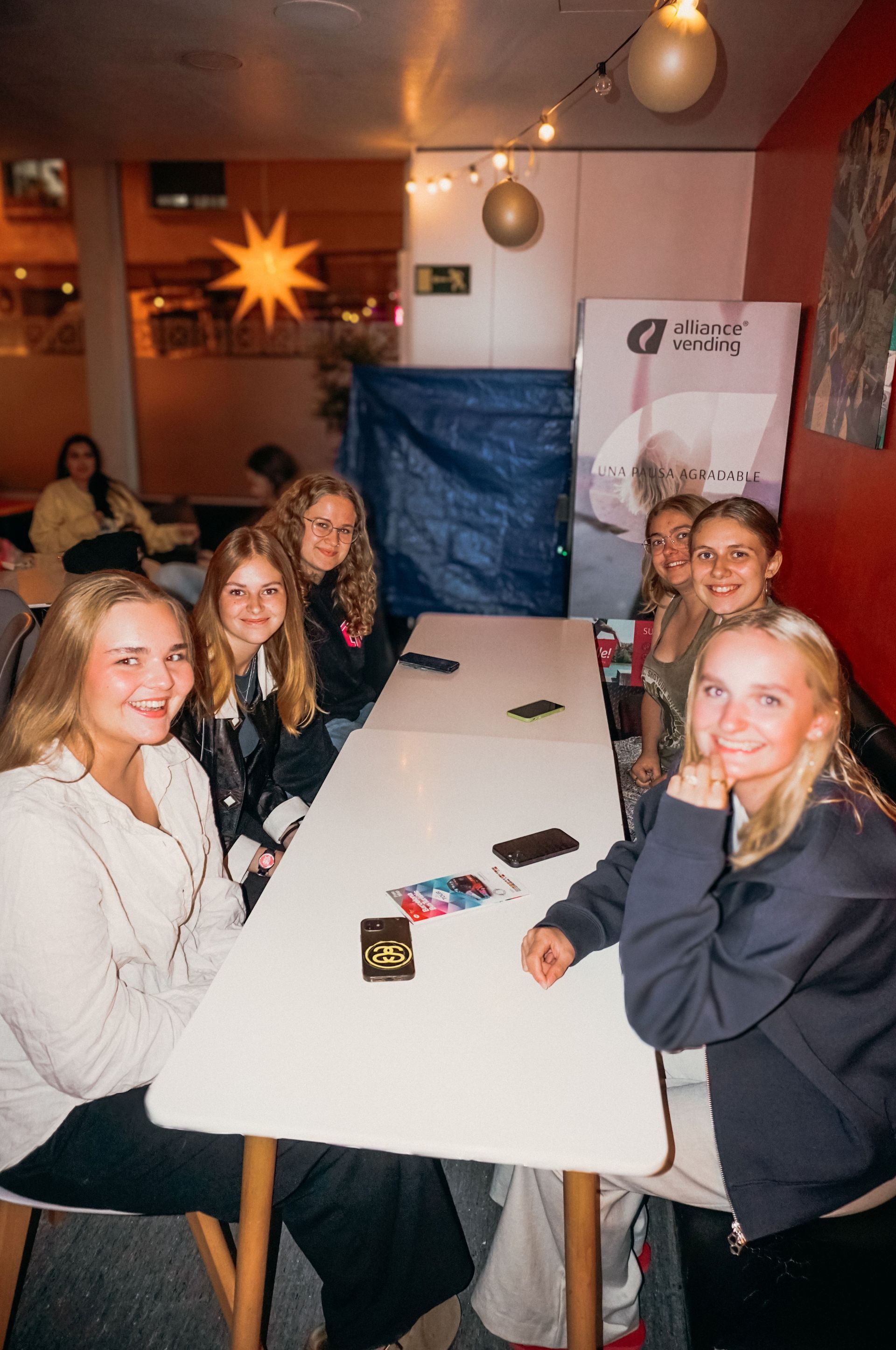 Un grupo de jóvenes reunidos alrededor de una mesa blanca en el interior, sonriendo y mirando a la cámara.