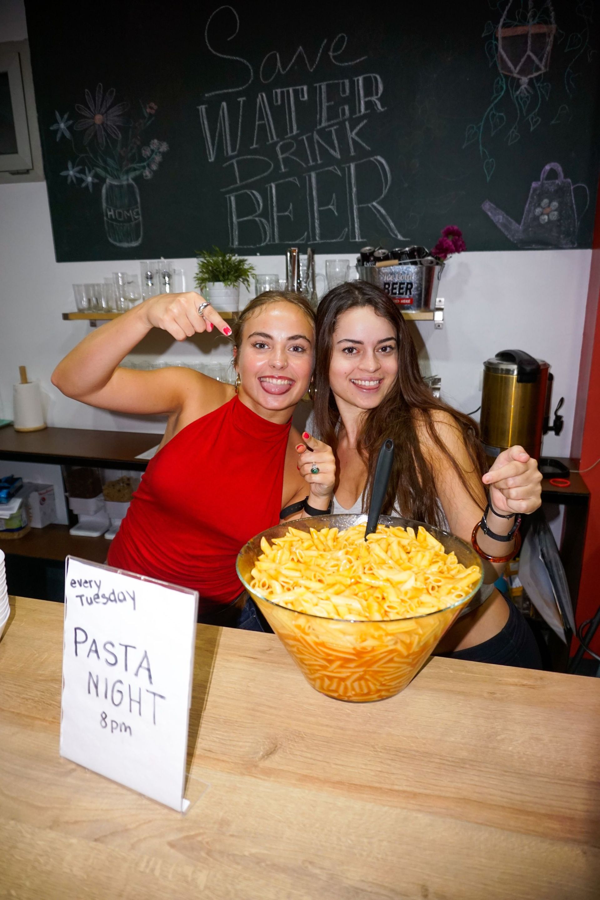 Dos mujeres posan junto a un gran plato de pasta, señalando. Cartel de 