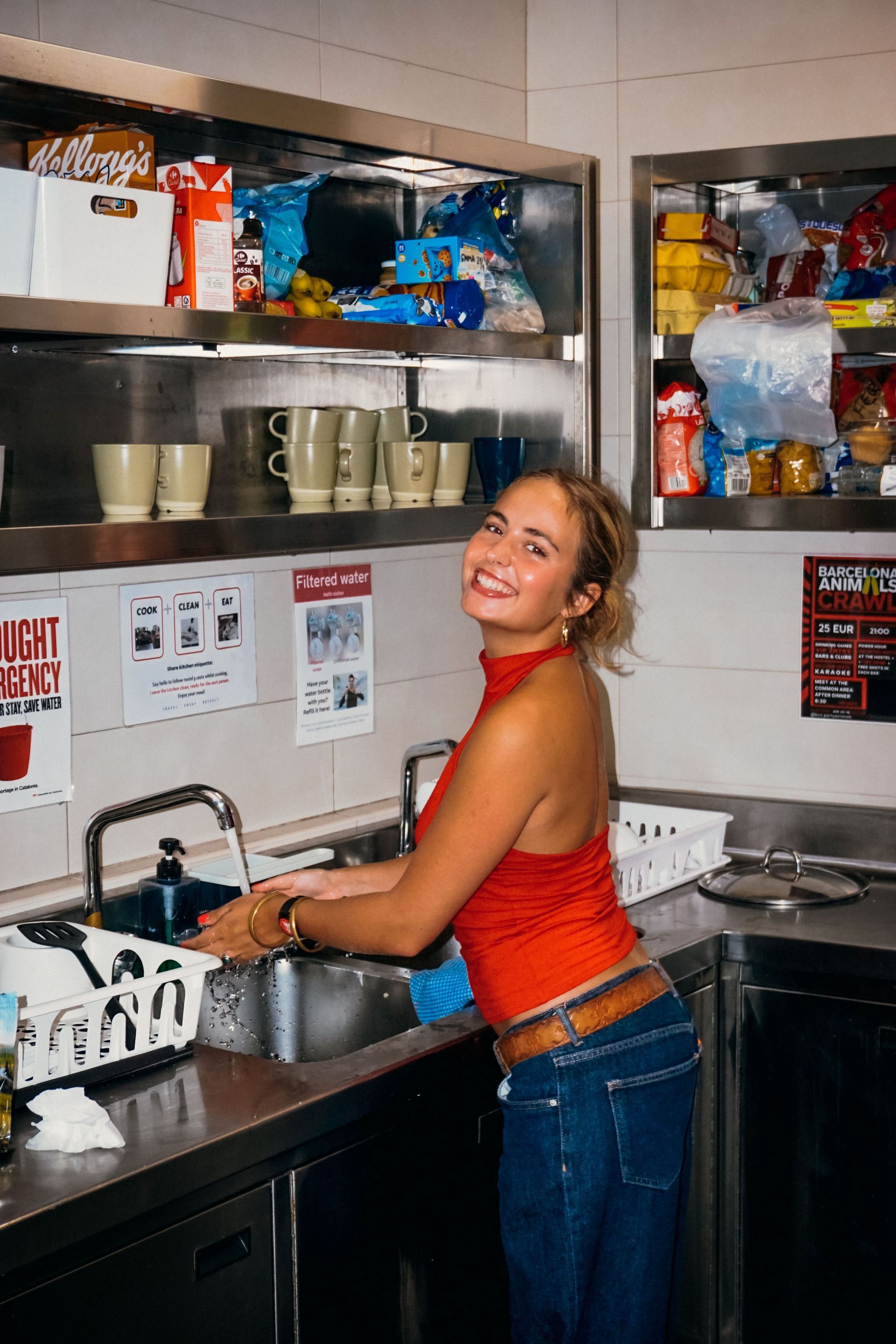 Cliente lavando platos en una cocina industrial, sonriendo. Mostradores de acero inoxidable, estantes con suministros. 