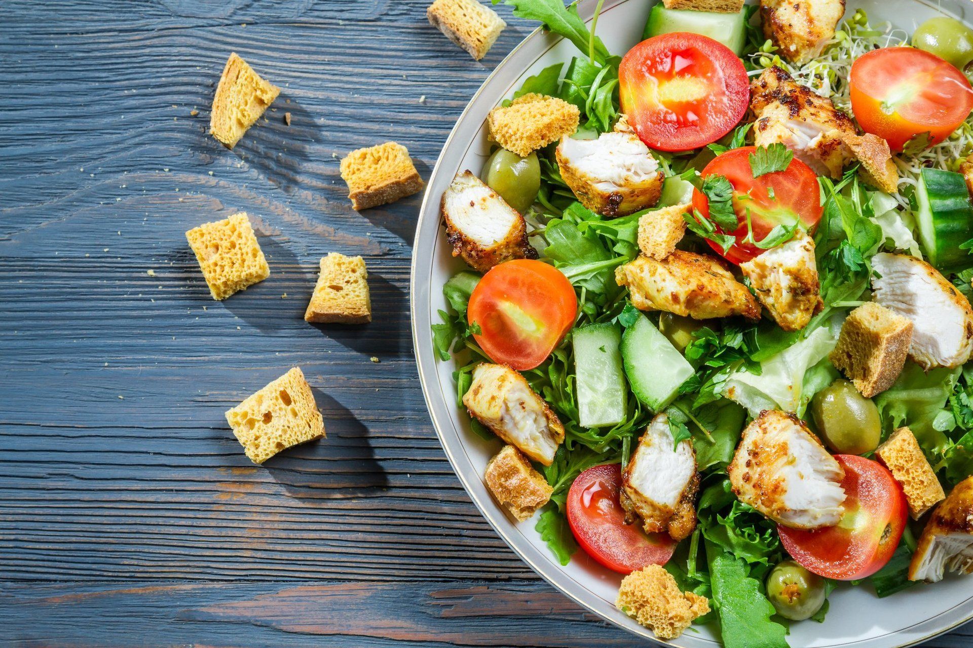 Chicken salad with croutons, tomatoes, and cucumbers in a bowl on a blue wooden surface.