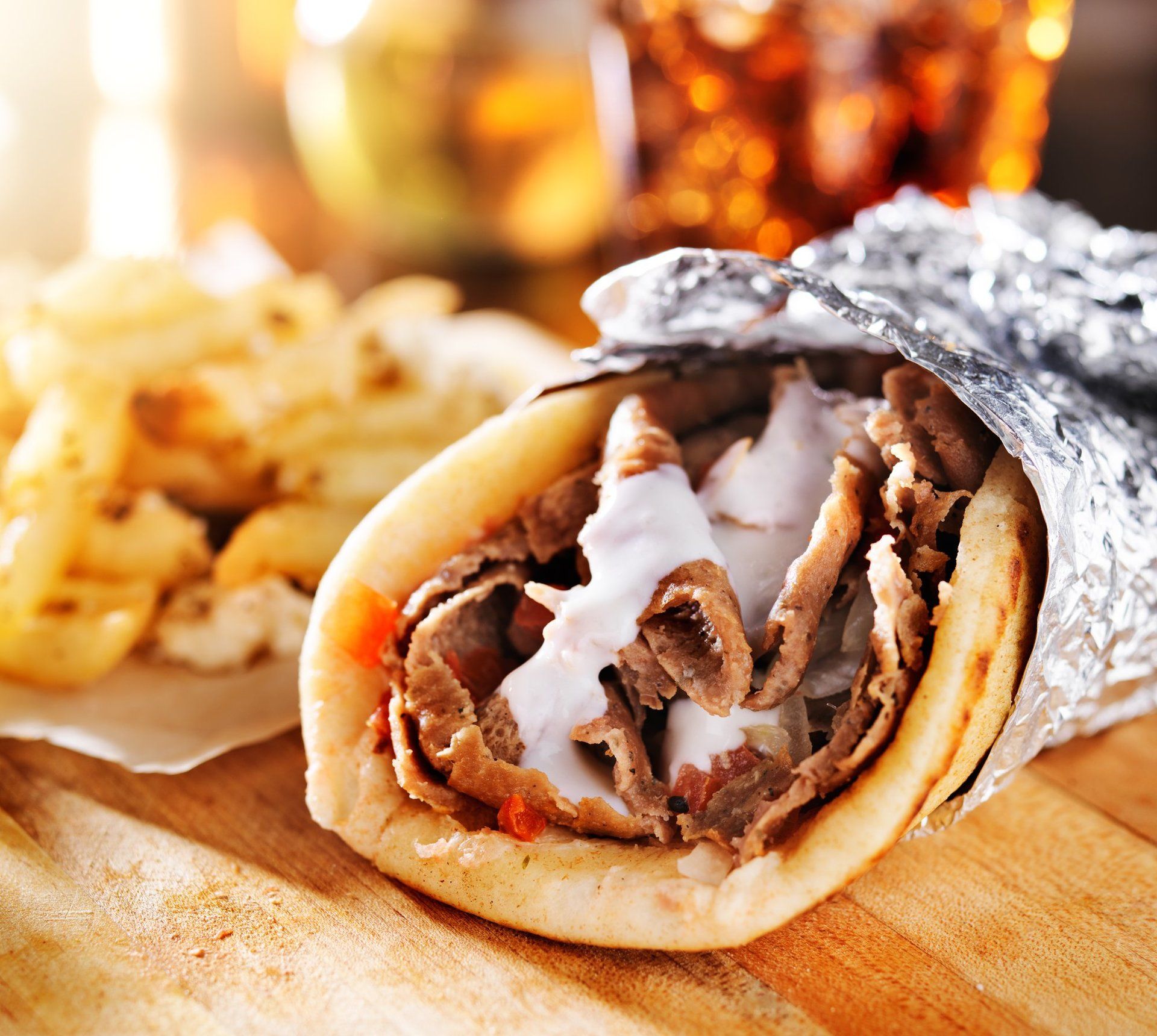 Gyro sandwich in foil with fries on wooden surface.