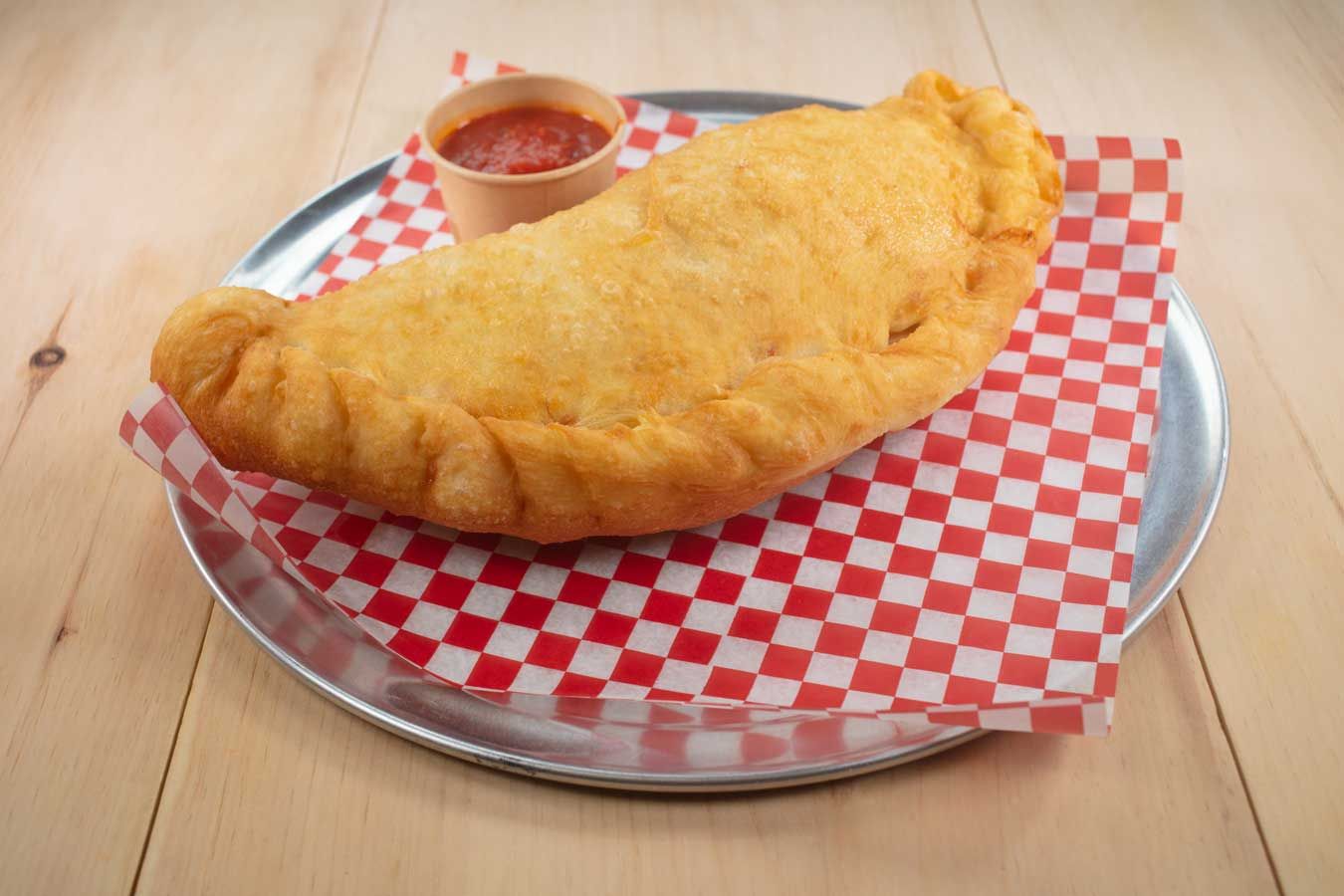 Fried calzone on a red and white checkered paper on a metal plate, with a small cup of sauce.