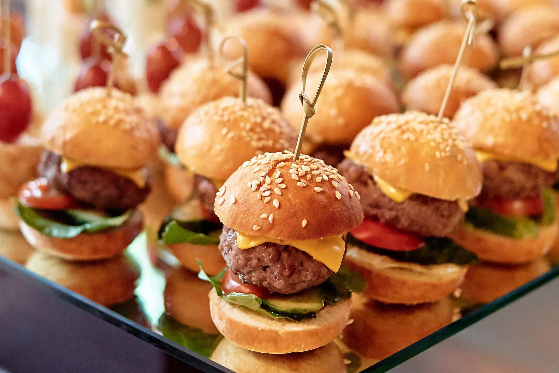 A bunch of mini hamburgers on toothpicks on a glass tray.