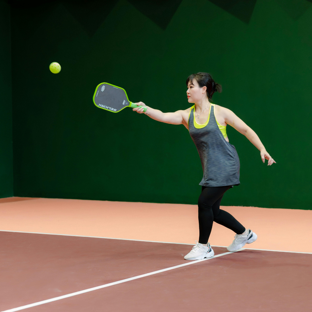 Woman playing pickleball, reaching for ball with green paddle on a court.