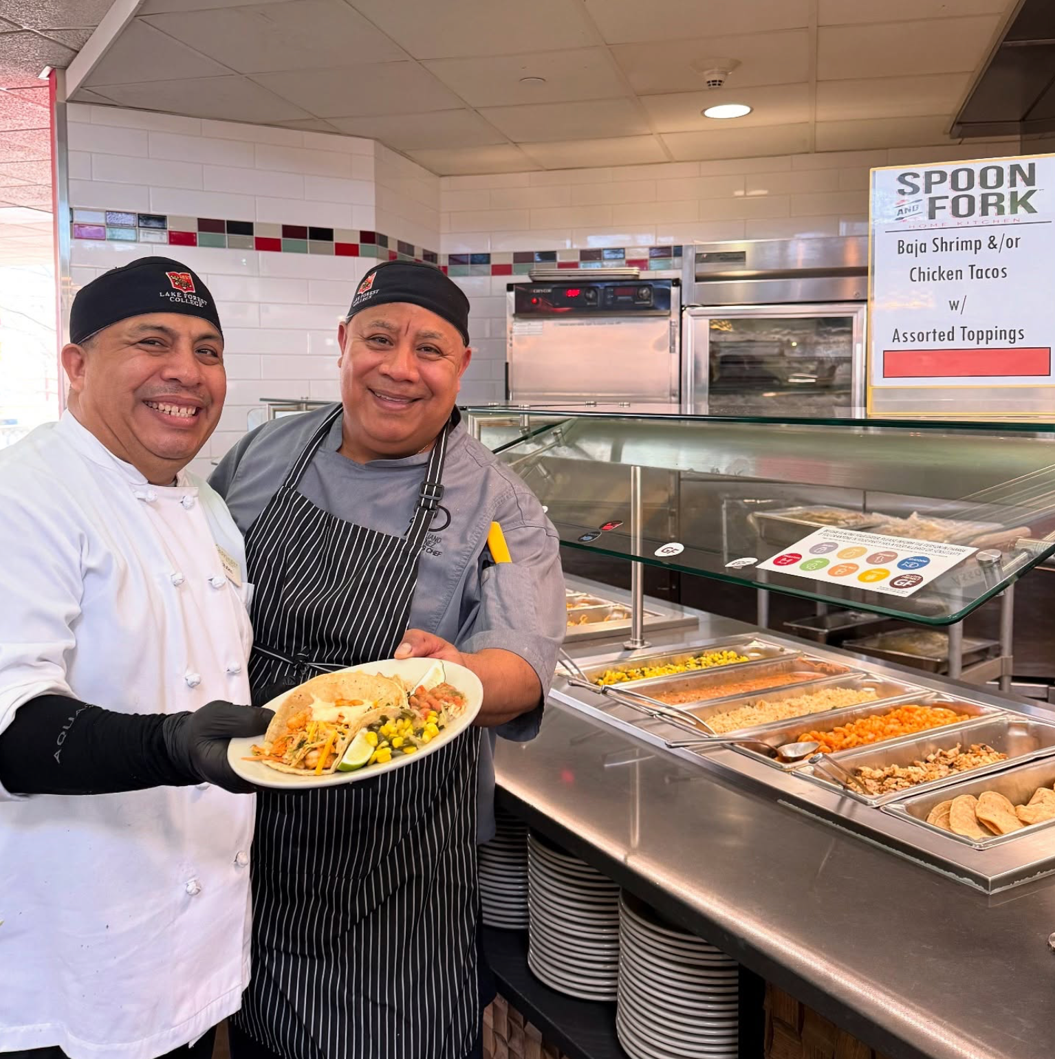 Two chefs smiling at a cafeteria counter, one holding a plate of food.