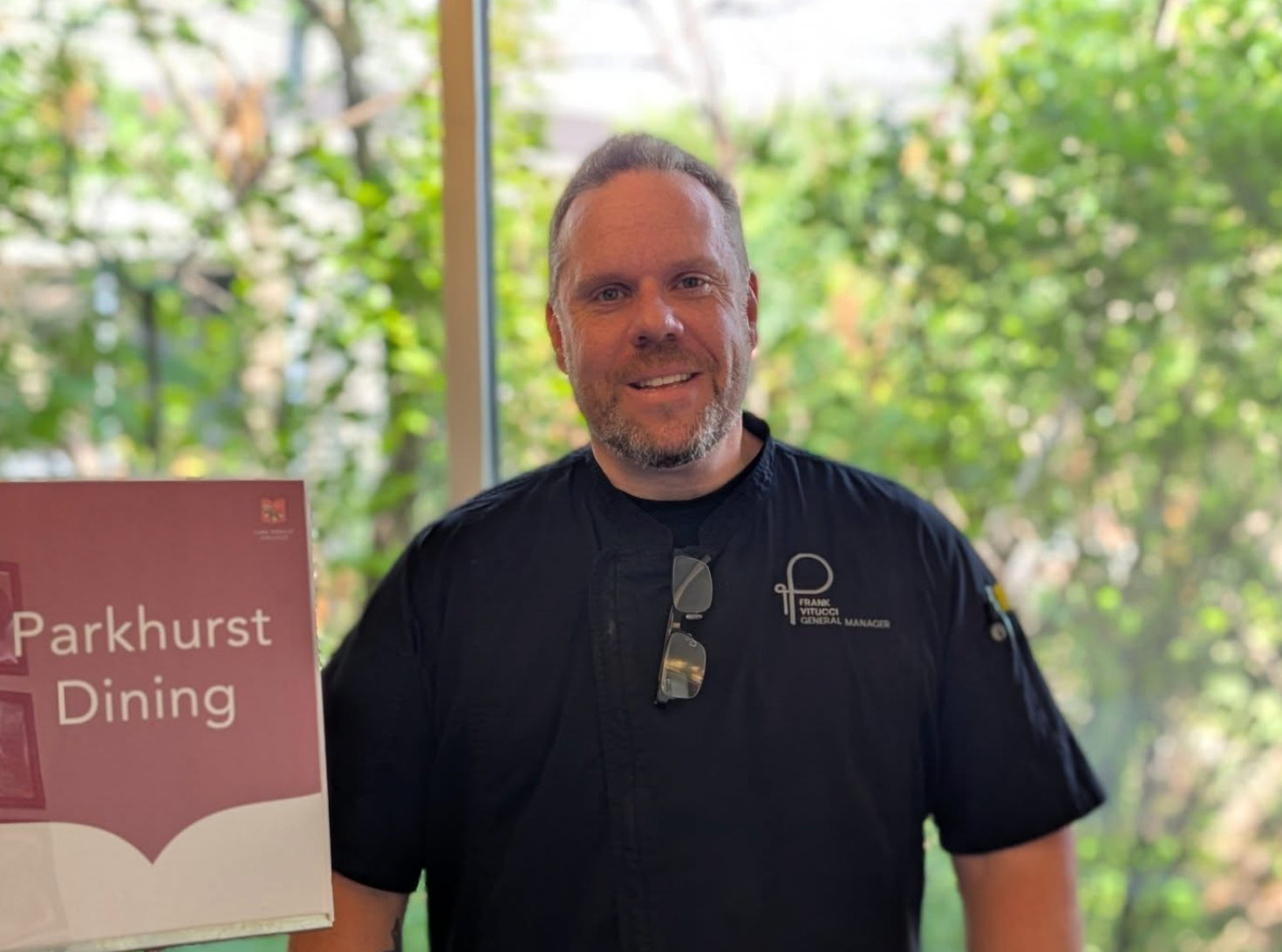 Man in black uniform smiles in front of Parkhurst Dining sign, near a window with greenery.