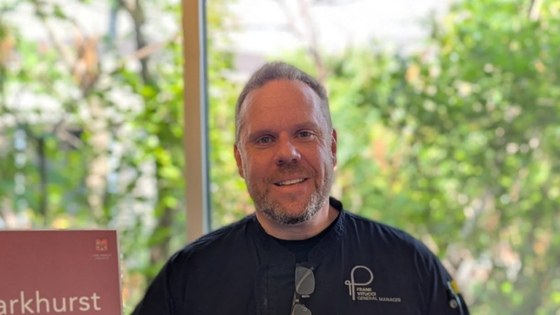 Man in black uniform smiles in front of Parkhurst Dining sign, near a window with greenery.