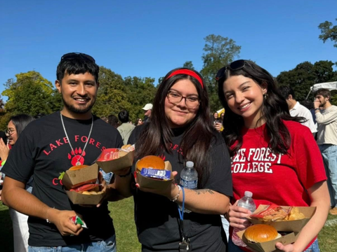 Three students hold boxed meals and smile outdoors on a sunny day.