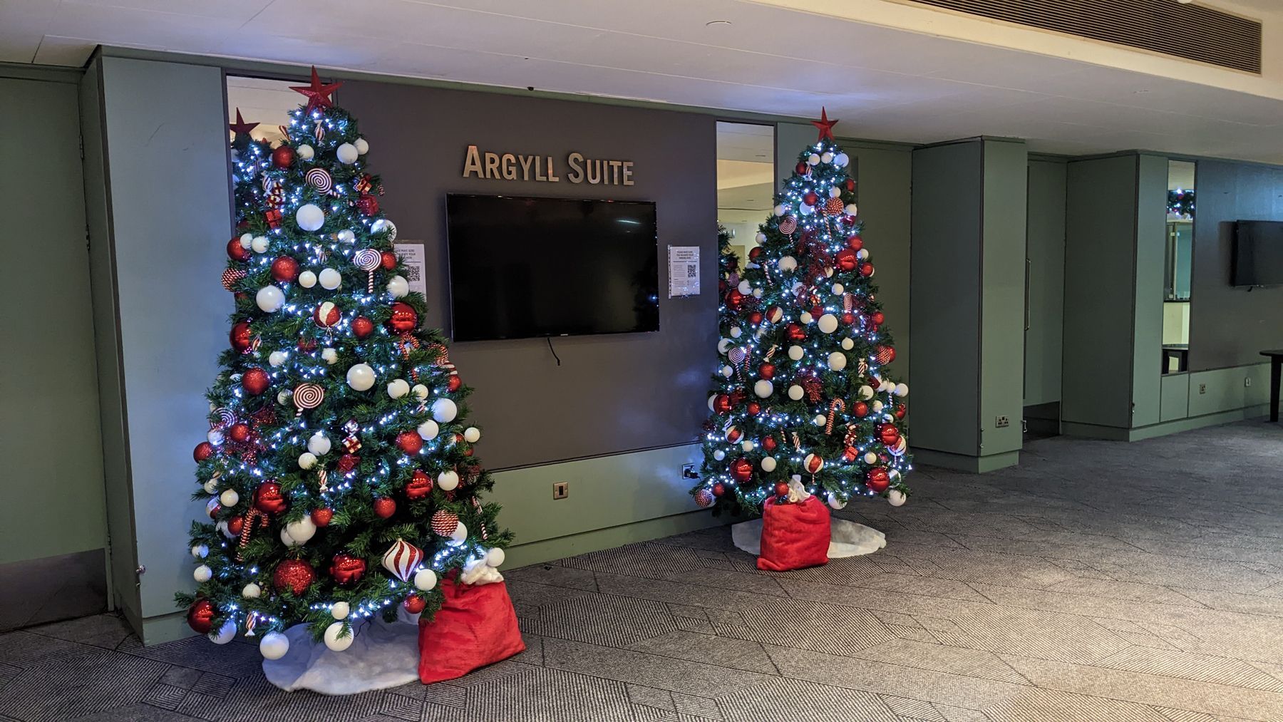 Two christmas trees are sitting in front of a wall in a hallway.