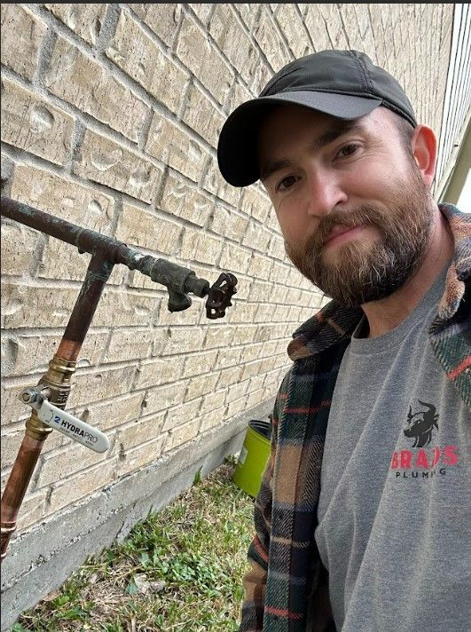 A man with a beard is standing in front of a brick wall next to a pipe.