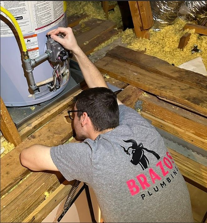 A man wearing a brazos plumbing shirt is working on a water heater