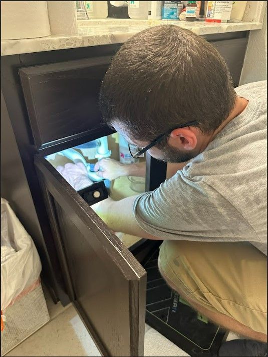 A man is kneeling down in a bathroom looking under a sink.