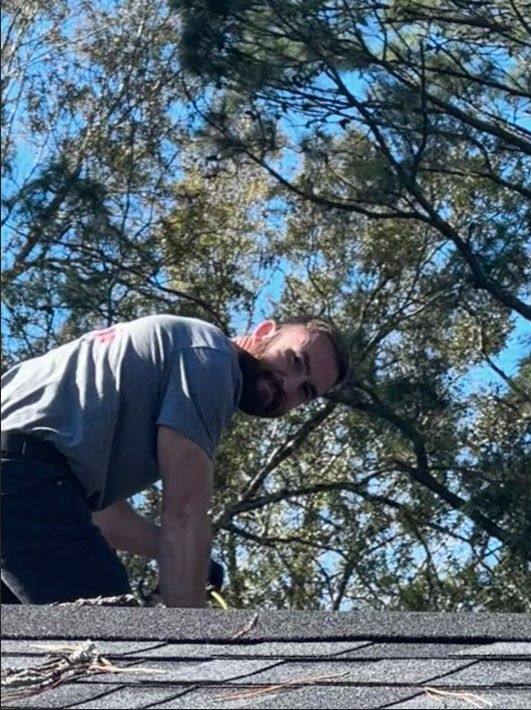 A man is kneeling on top of a roof with trees in the background.