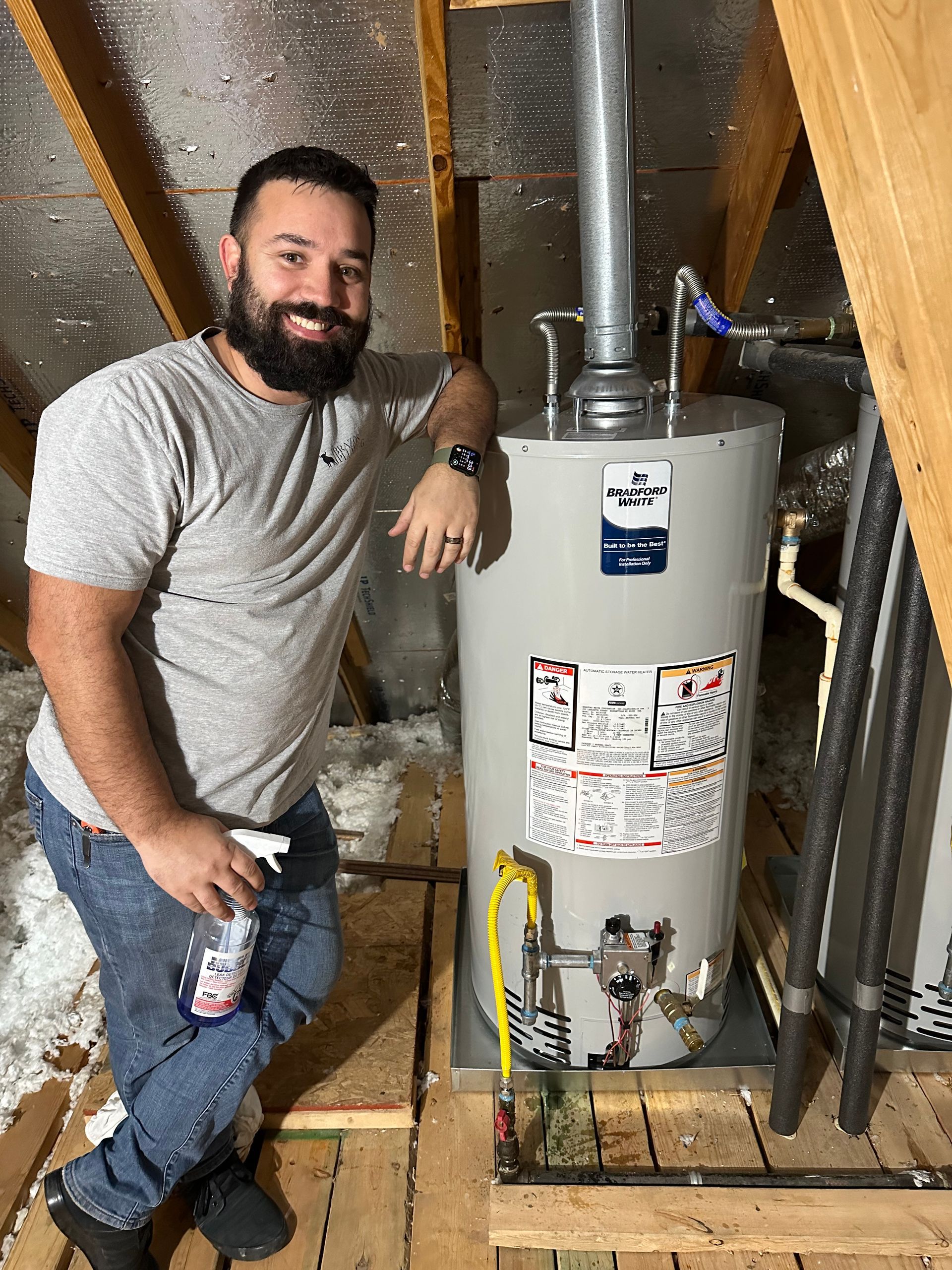 A man with a beard is standing next to a water heater in a attic.