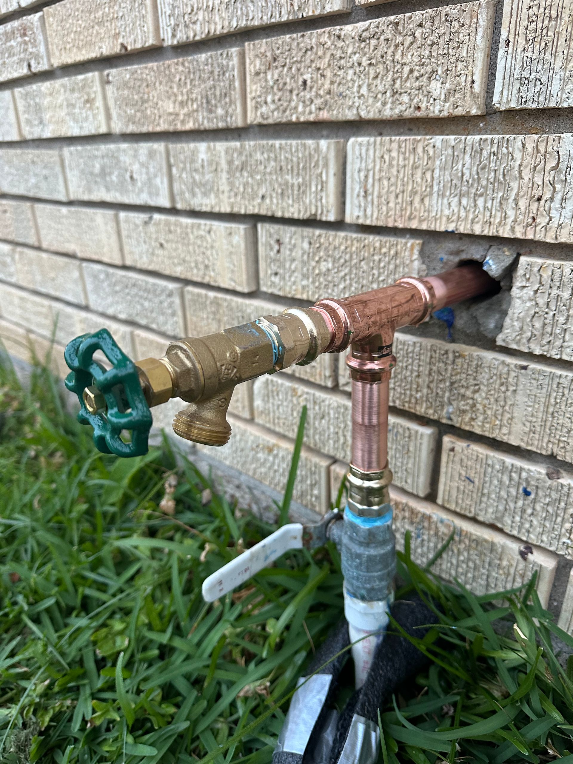 A faucet is connected to a brick wall and is sitting in the grass.
