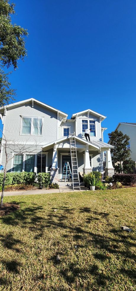 A light gray two-story house with a white porch, green landscaping, and a person on the roof under a clear blue sky.
