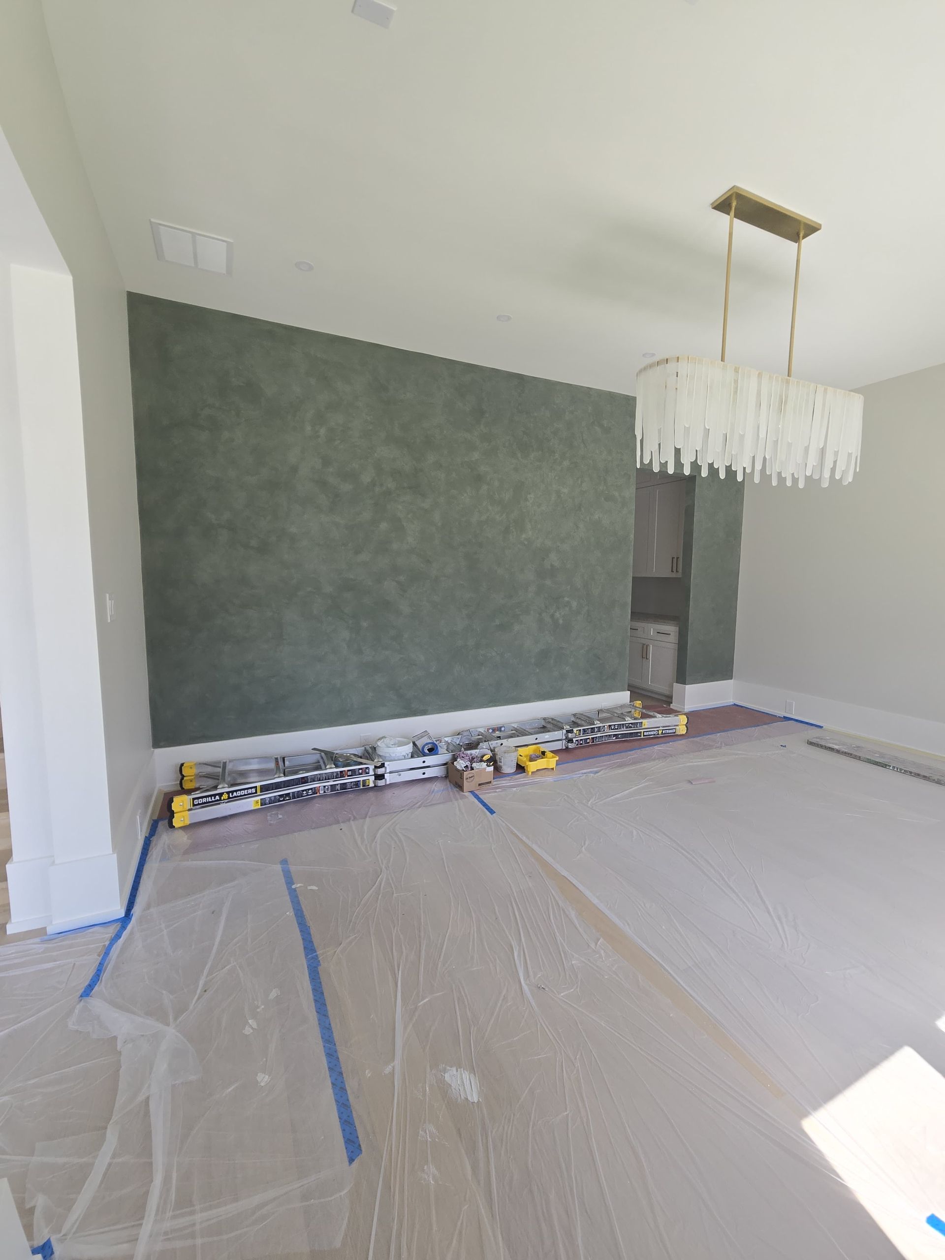 A room under construction with a large, textured, moss-green accent wall and a modern, tiered crystal chandelier above.
