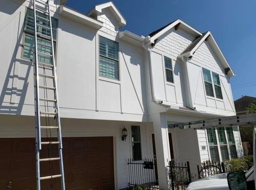 An extension ladder leans against the side of a two-story white suburban house with a brown garage door.