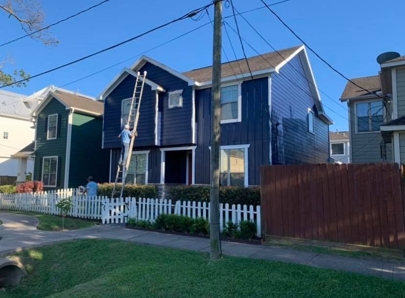 A worker on a ladder paints the dark blue exterior of a two-story house behind a white picket fence.