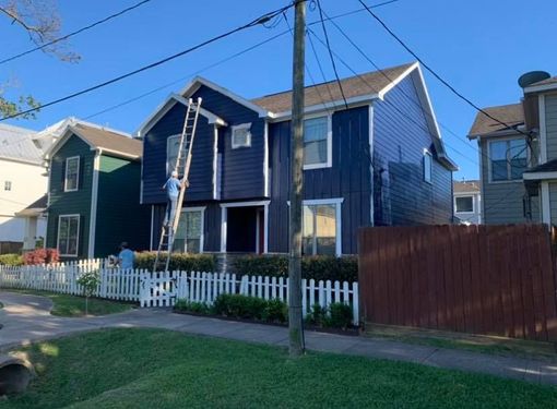 A worker on a ladder paints the dark blue exterior of a two-story house behind a white picket fence.