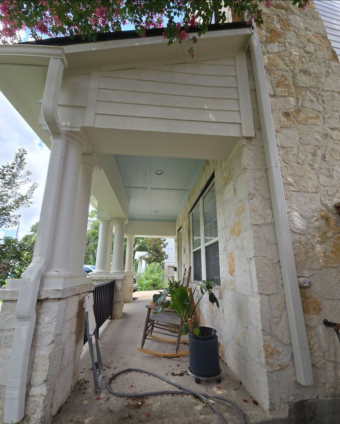 A covered porch with a stone wall, white columns, blue ceiling, and a wooden rocking chair.