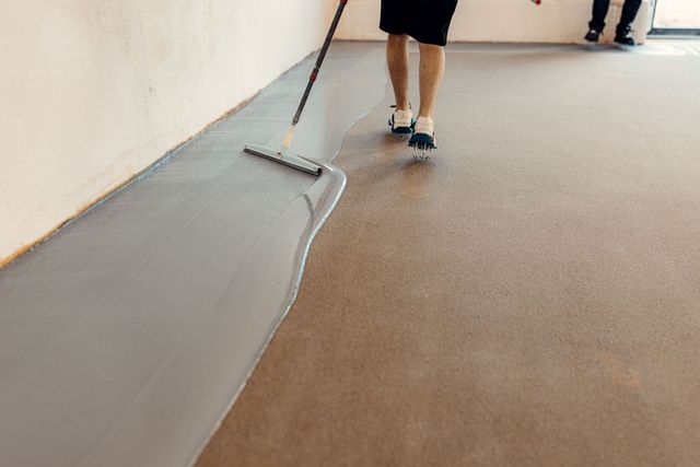 Person spreading gray floor coating with a squeegee beside a brown floor in a hallway