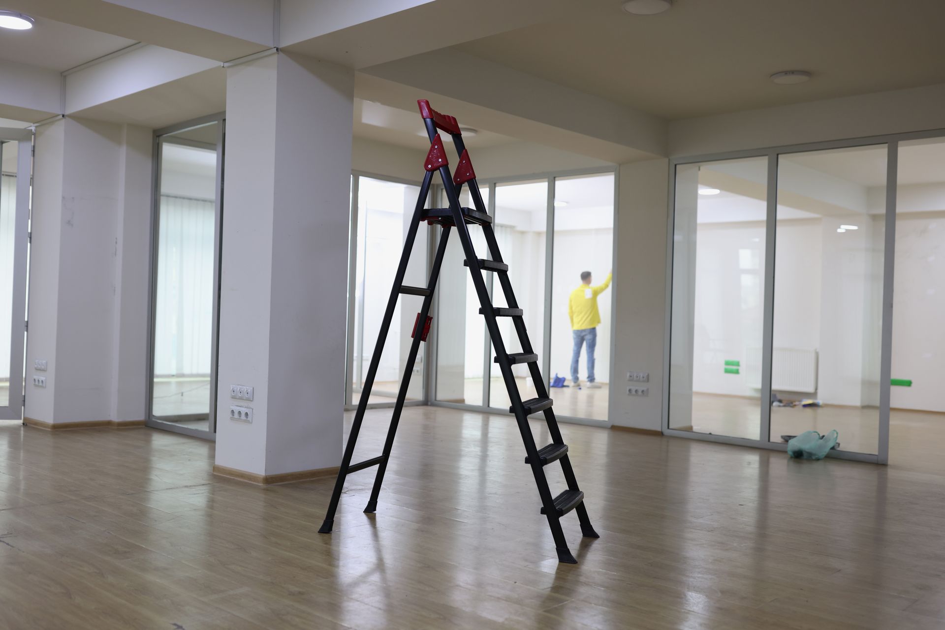 A black stepladder stands in a bright, empty room with glass partitions while a person works in the background.