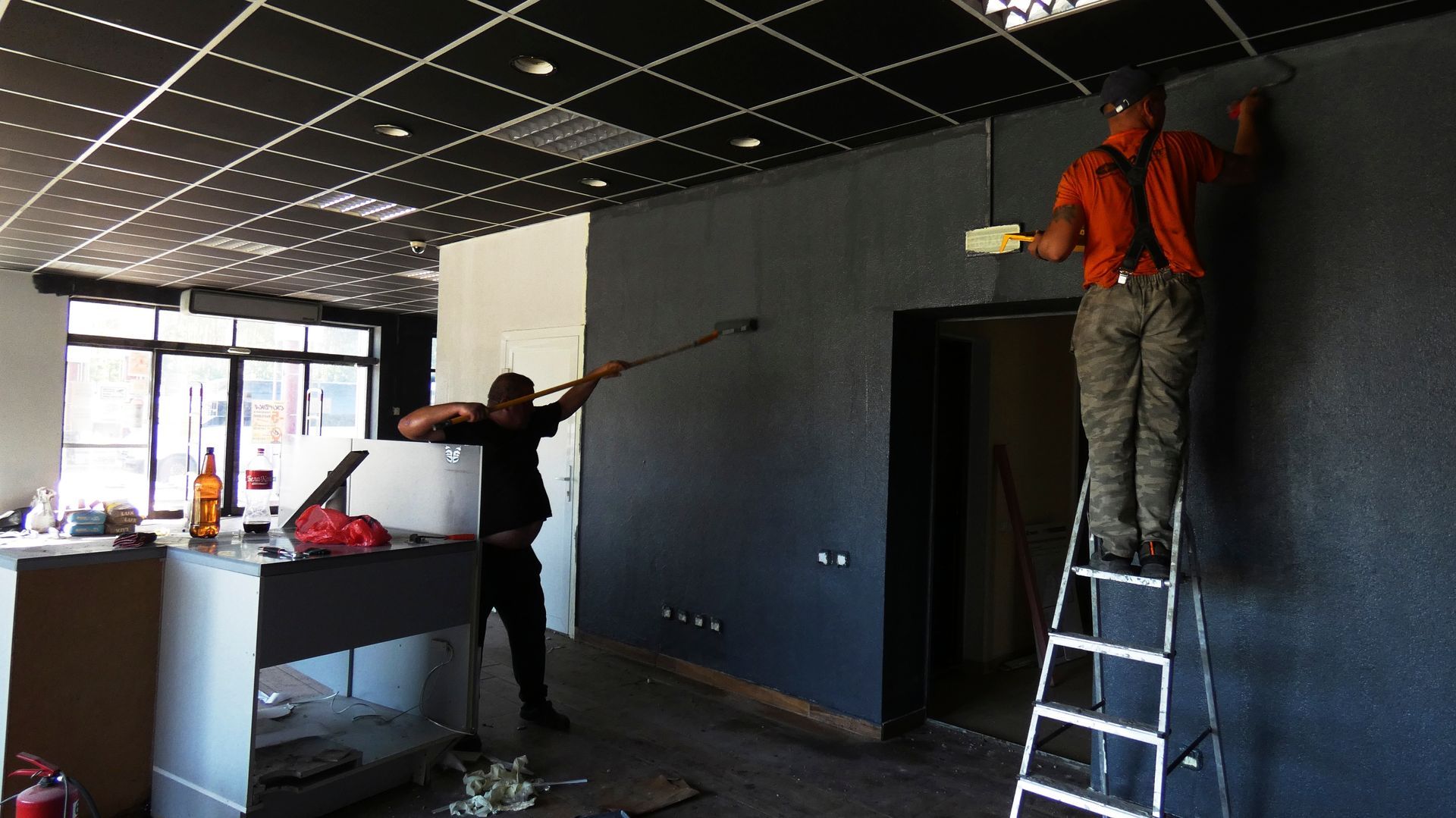 Two construction workers paint a dark wall in an unfinished indoor space with a black grid ceiling.