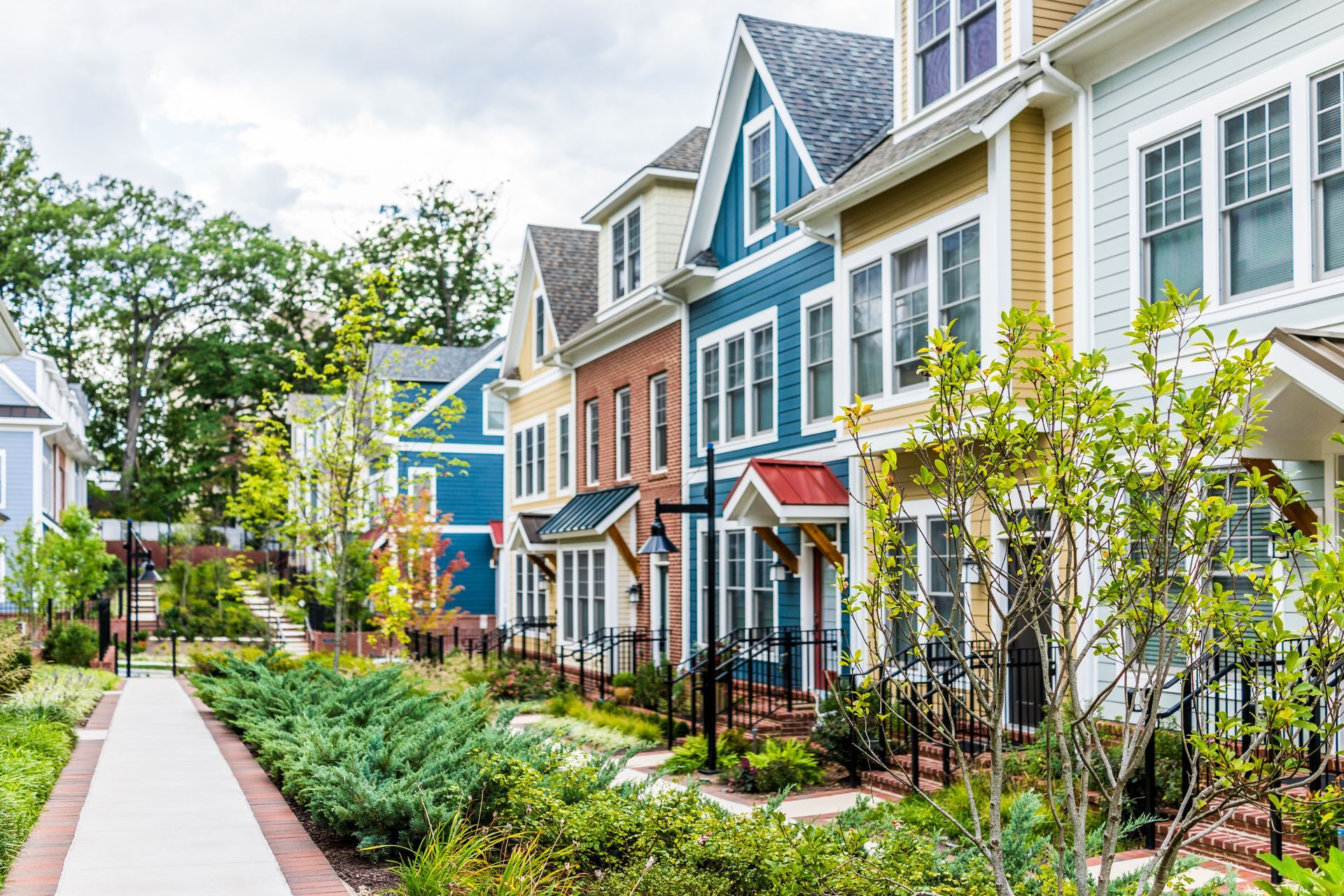 A row of colorful townhouses featuring varying siding colors, pitched roofs, and a paved walkway lined with green bushes.