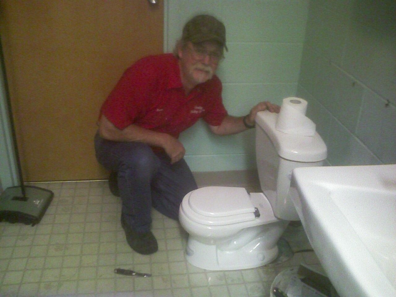 A man in a red shirt is squatting next to a toilet