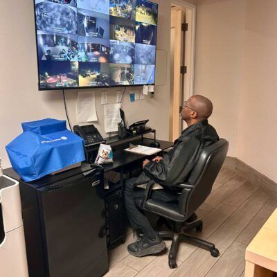 Man sits at desk watching security monitors. Office setting, monitors display various camera feeds.