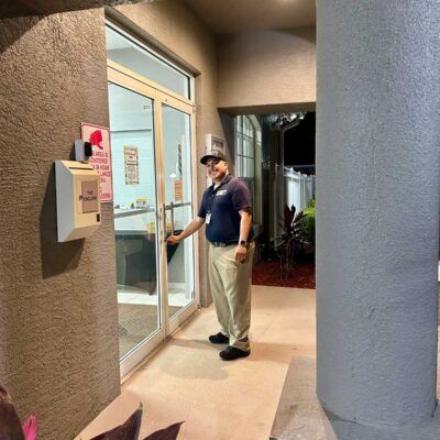 Man in uniform opening glass door. Outside, neutral-toned building with mailbox, lit at night.