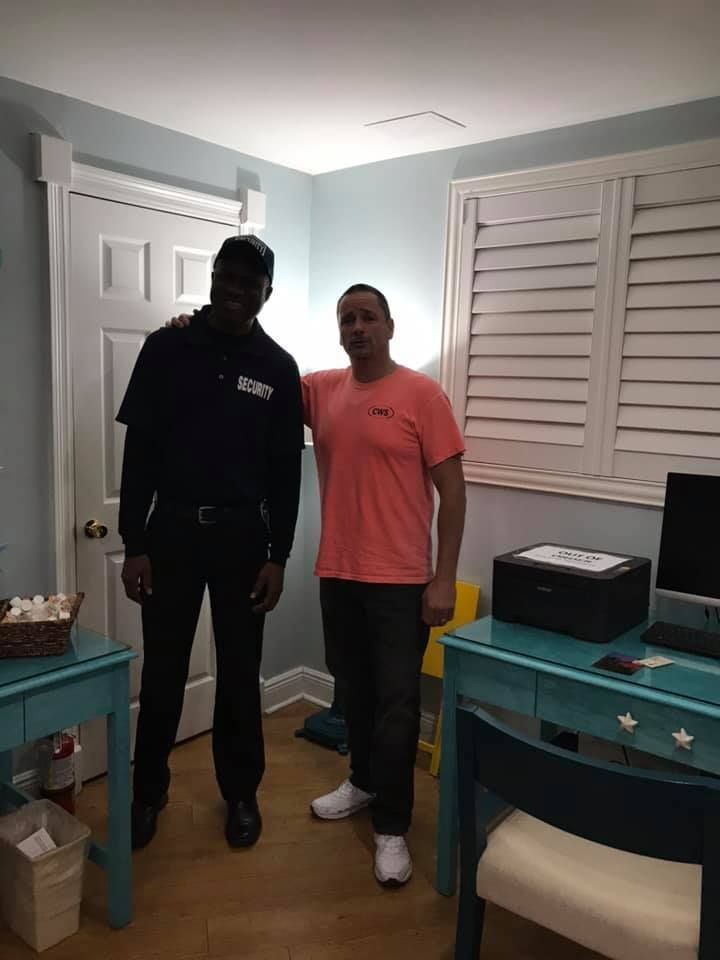 Two men stand indoors. One in dark uniform, the other in orange shirt. They are in a room with blue desks and white shutters.