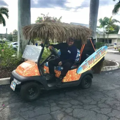 Person in decorated golf cart with surfboard. Palm trees in background.