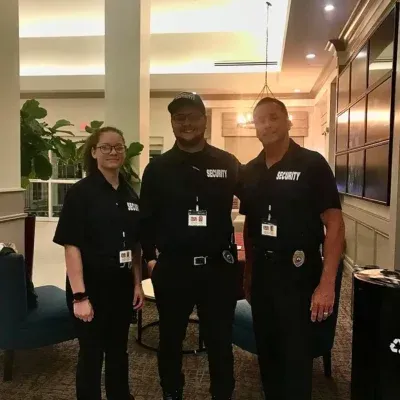 Three security guards posing together, wearing black uniforms, inside a building with a lobby area.