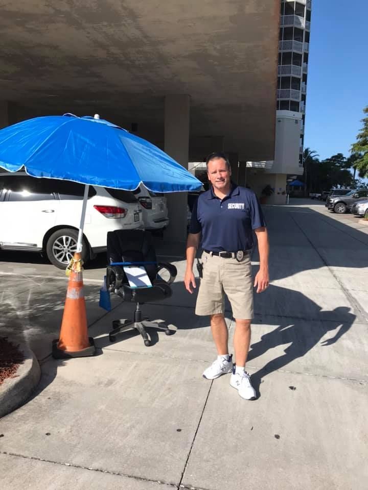 Man in uniform stands by an office chair under a blue umbrella near a white SUV and a tall building.