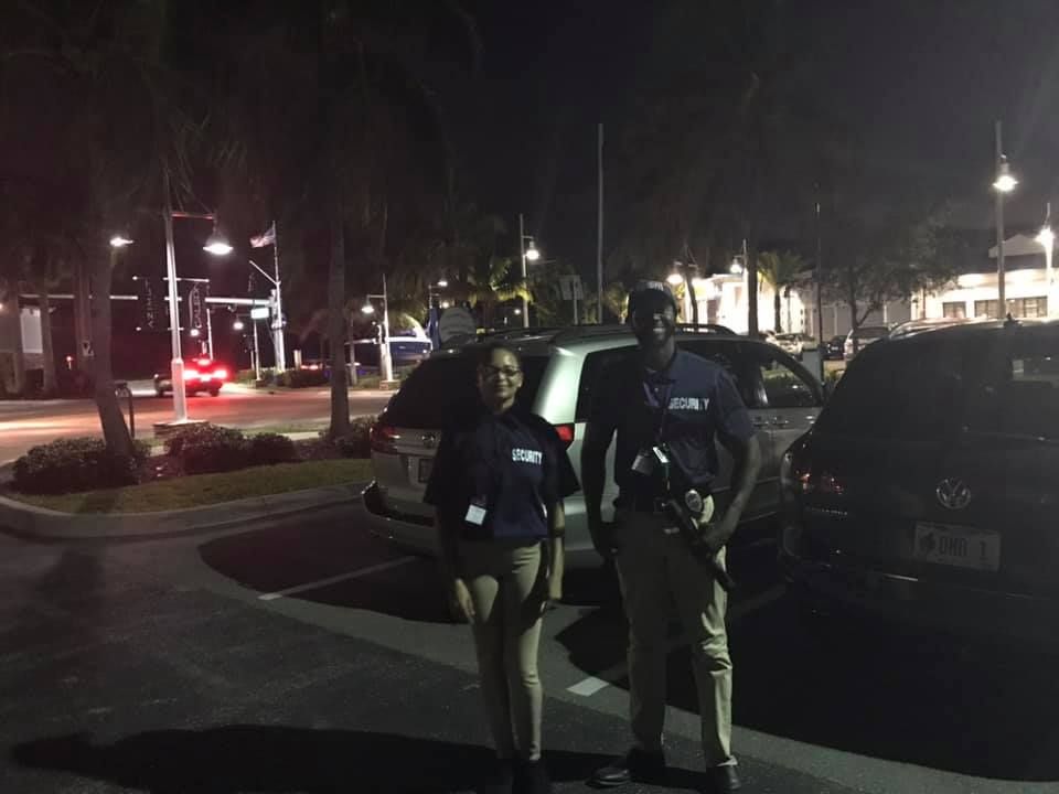 Two security guards standing in a parking lot at night; one holding a rifle.