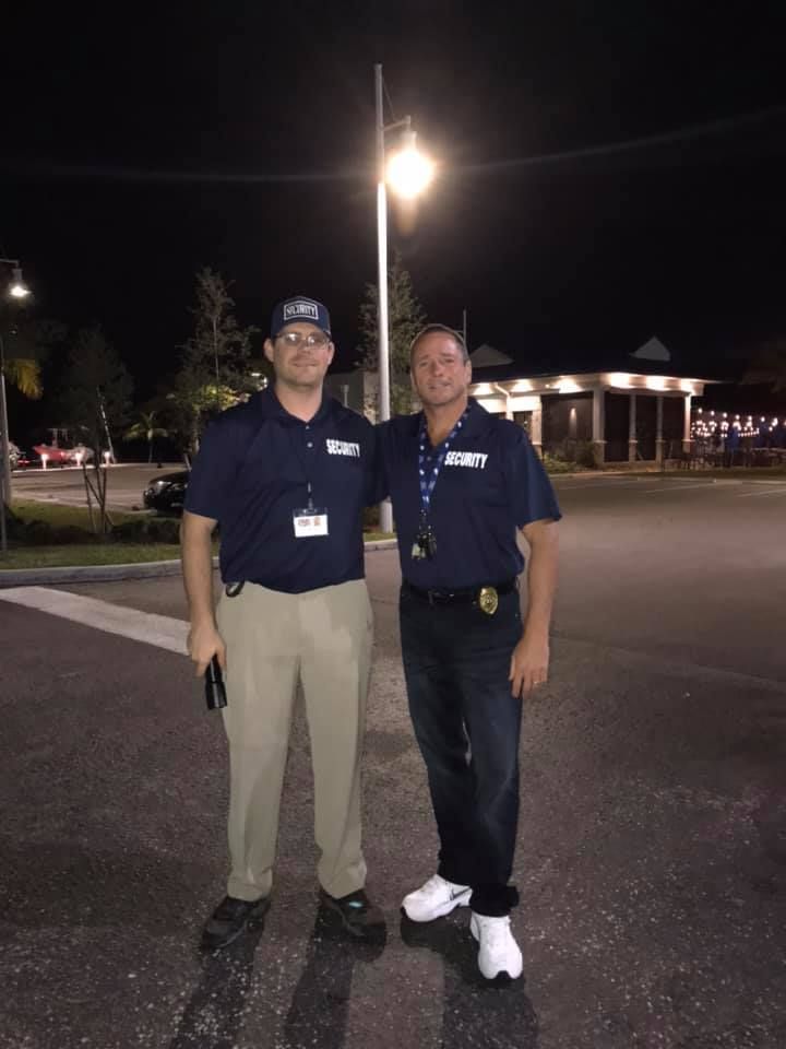 Two men in security uniforms pose outside at night under a streetlight. One wears tan pants, the other wears jeans.